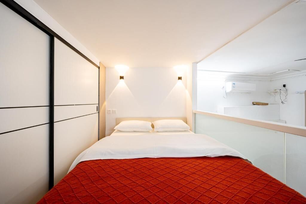 Ein Hotelzimmer mit einem Bett, weißen Wänden und einer roten gesteppten Tagesdecke. Zwei Wandlampen sind über dem Bett angebracht, und auf der rechten Seite befindet sich ein kleines Fenster.