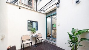 Un petit espace patio avec deux chaises et une table, une plante en pot et une fenêtre donnant sur un bâtiment situé de l’autre côté de la rue.