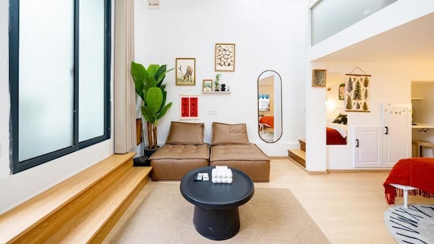 Un salon moderne avec un canapé, une table basse et un grand miroir. On aperçoit, au fond, une plante, un fauteuil rouge et un coin cuisine.