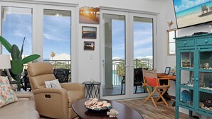 Ein Wohnzimmer mit einem beigefarbenen Sofa, einem Couchtisch mit Meeresschalen, einem türkisblauen Schrank und einem Fernseher, der eine Strandansicht zeigt.