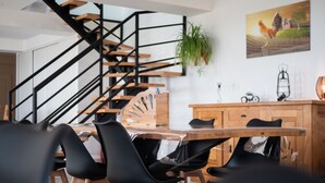 Un comedor moderno con una mesa de madera, sillas negras, una escalera de madera y una cocina al fondo.