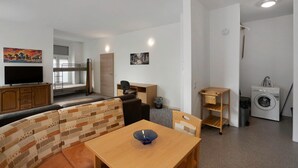 A compact living space with a sofa, a dining table, a TV, a washing machine, and a small kitchen area.