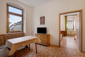 A room with a table, a TV on a wooden cabinet, a bench, and a view of buildings through a window.