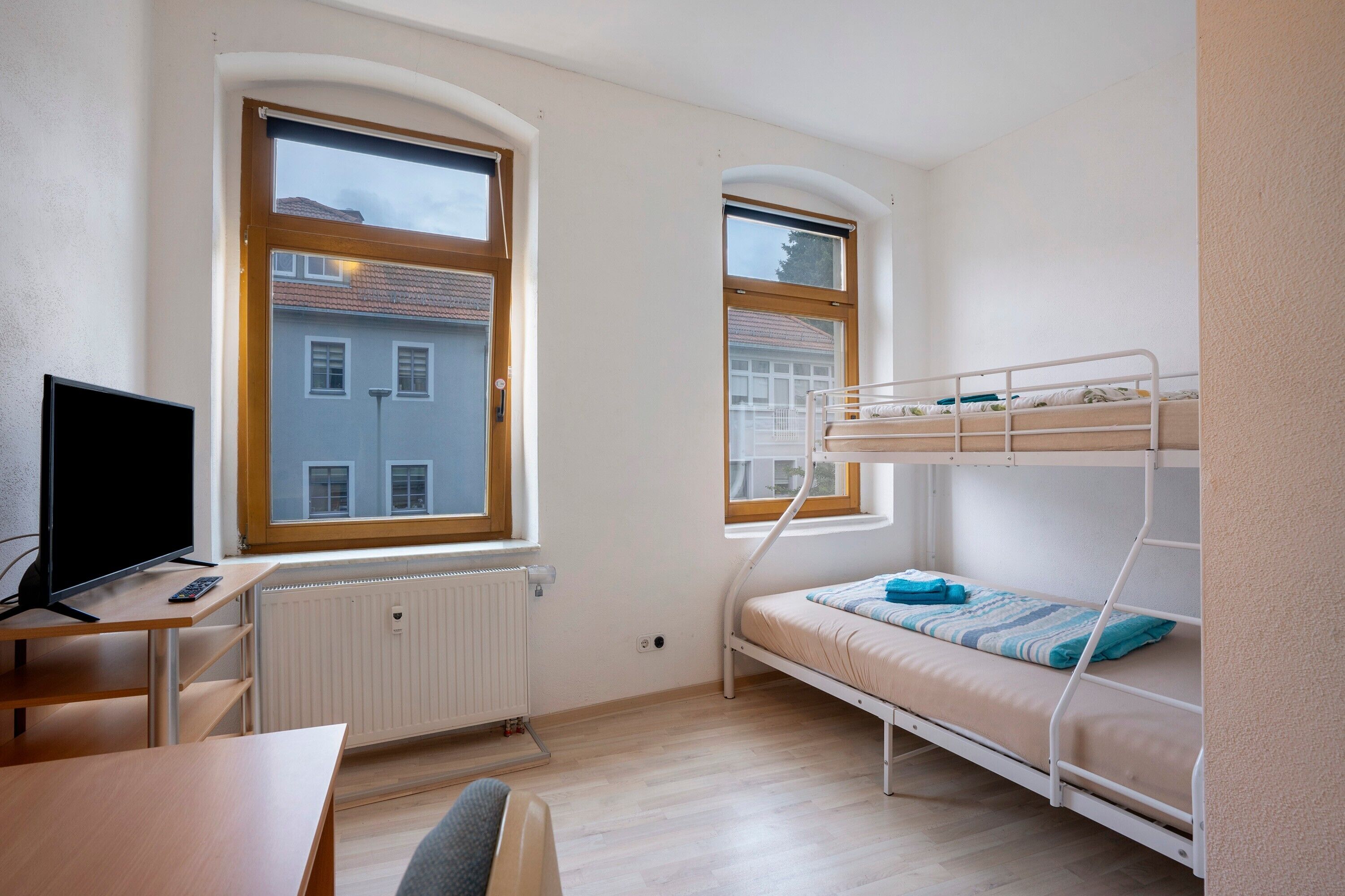 Ein Zimmer mit einem Etagenbett, einem Schreibtisch mit Computer, einem Fenster und einer Heizung.