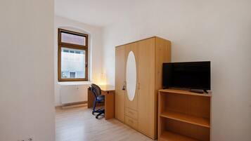 Ein Zimmer mit Holzschrank, Fernseher auf einem Regal, Schreibtisch mit Stuhl und Fenster mit Jalousien.
