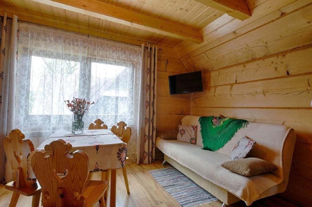 A wooden cabin interior with a dining table, chairs, a sofa, and a flat-screen TV mounted on the wall.