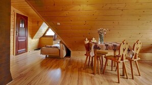 Een gezellig zolderkamertje met houten wanden en een schuine hemel. Er staat een houten tafel met stoelen, een bankje en een vaas met bloemen. Een deur en een raam zijn zichtbaar.