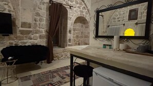 Ein rustikaler Raum mit Steinwänden, ausgestattet mit einem Bett, einem kleinen Tisch, einem Barhocker, einem Kühlschrank und einem Spiegel mit Bilderrahmen.