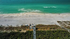 Vista aérea de una playa con aguas turquesas cristalinas, una orilla arenosa y algunas construcciones, incluyendo una escalera que baja a la playa.