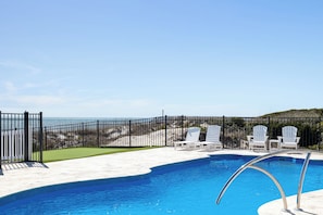 Une piscine avec vue sur la plage, entourée d’une clôture et de chaises longues blanches.