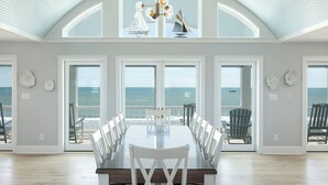 Uma sala de jantar ampla, com uma mesa grande e cadeiras, janelas grandes com vista para o oceano e teto alto com claraboia.