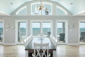 A spacious dining area with a large table and chairs, large windows with ocean views, and a high ceiling with a skylight. - Casa Blanco: Oceanfront House - 10 BR 11.5 BA (Emerald Isle)