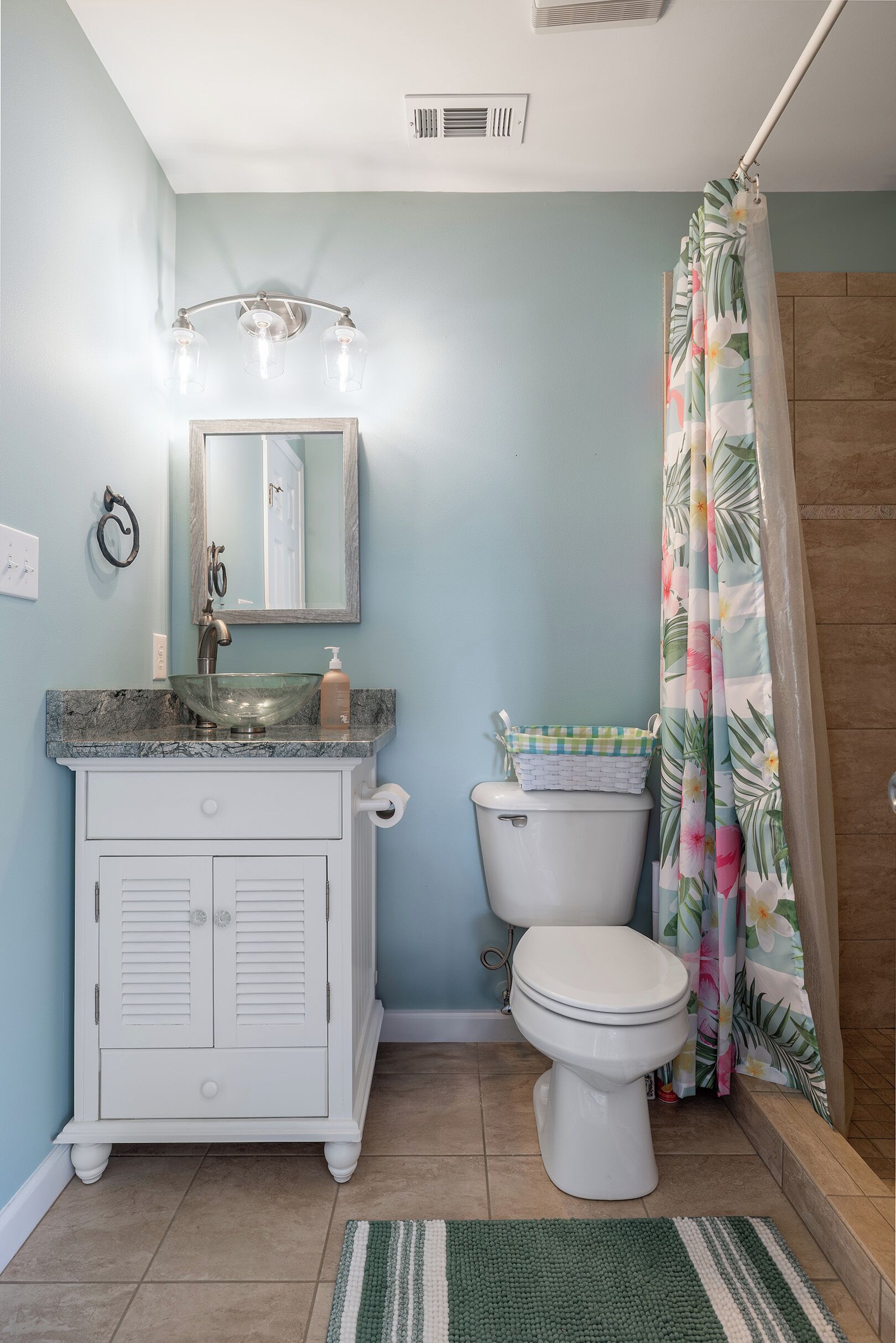 A bathroom with a marble sink, a toilet with a basket, a shower curtain with a tropical pattern, and a green and white striped rug.