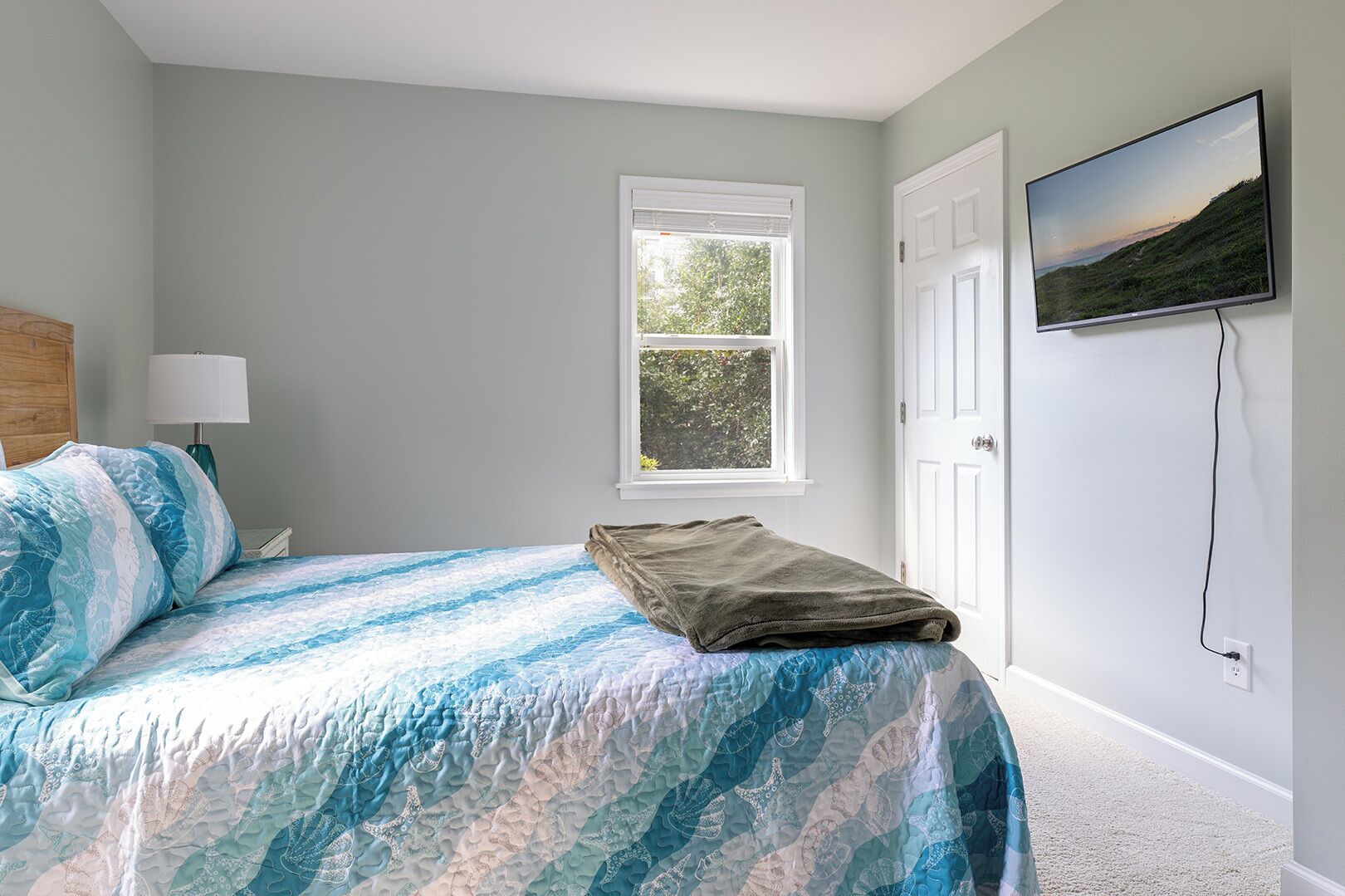 A bedroom with a bed, a window, a door, and a wall-mounted TV.