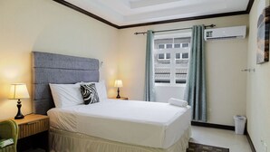 Une chambre d’hôtel avec un grand lit, deux tables de chevet avec des lampes, une fenêtre avec des rideaux, un appareil de climatisation et une poubelle.