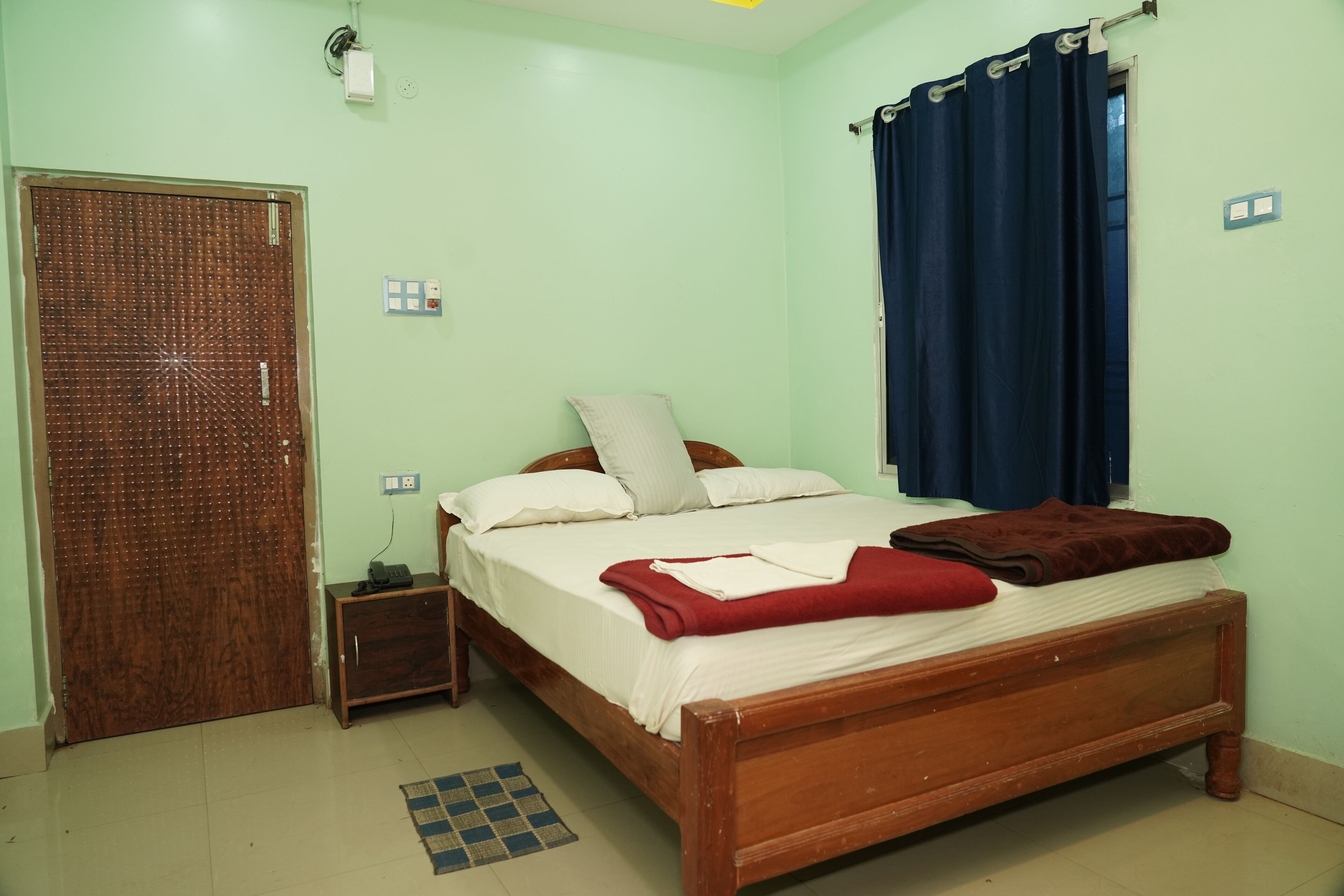 A bedroom with a wooden bed, a small bedside table, a wall socket, and a window with curtains.