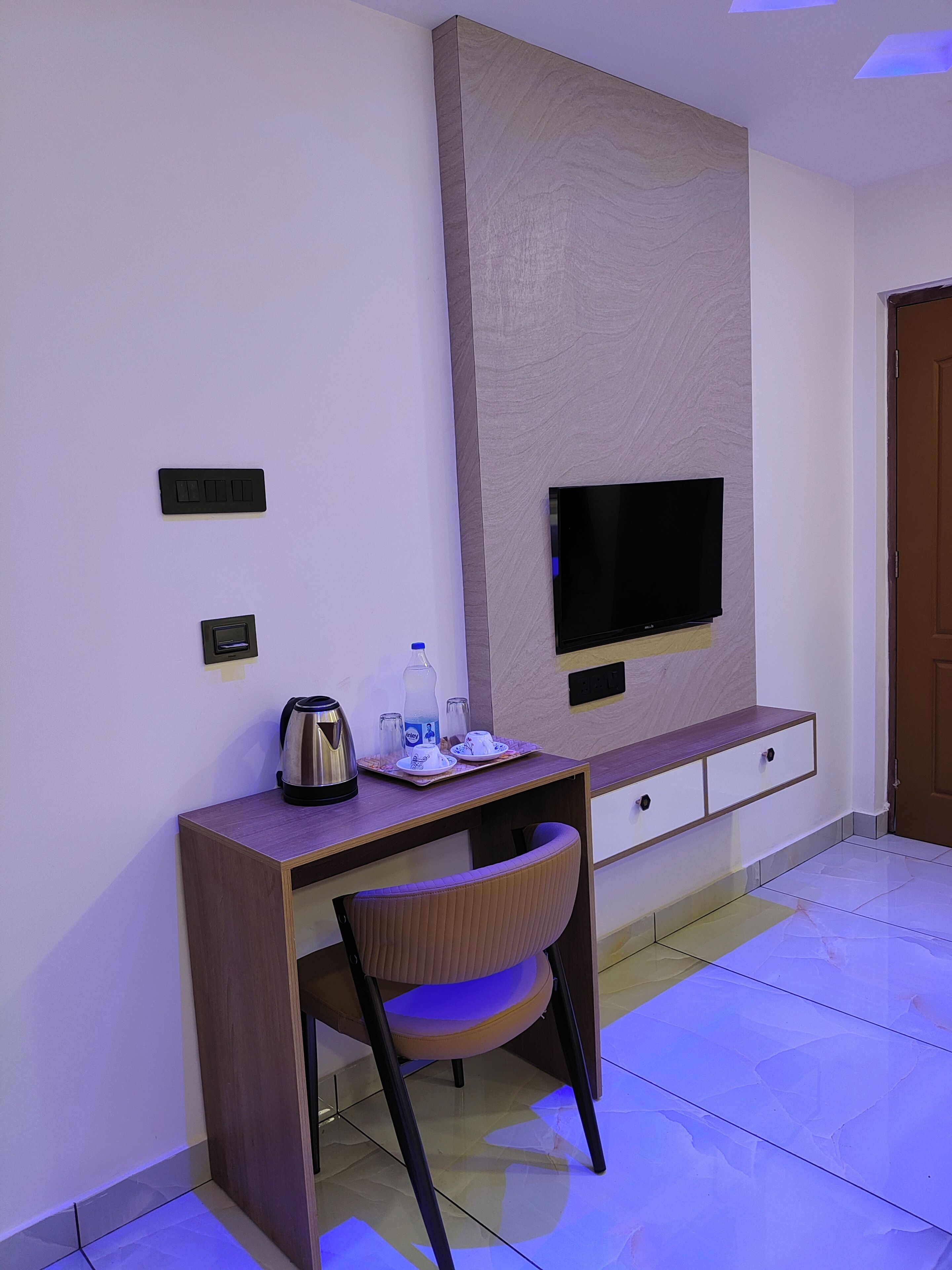 A modern hotel room with a wooden desk, a chair, a flat-screen TV mounted on the wall, and a small cabinet below.