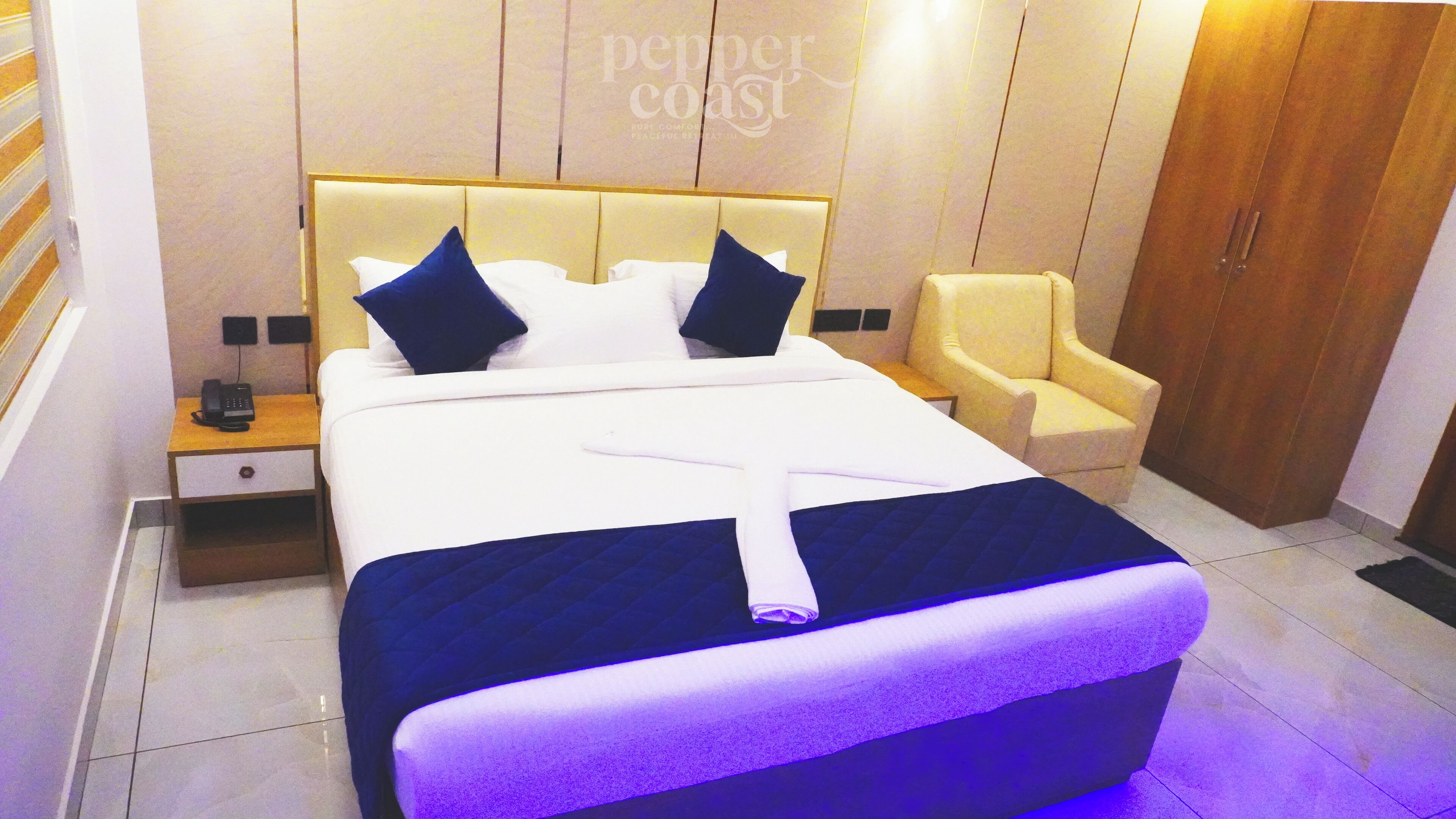 A modern hotel room with a large bed, a bedside table with a phone, a chair, and a wardrobe.