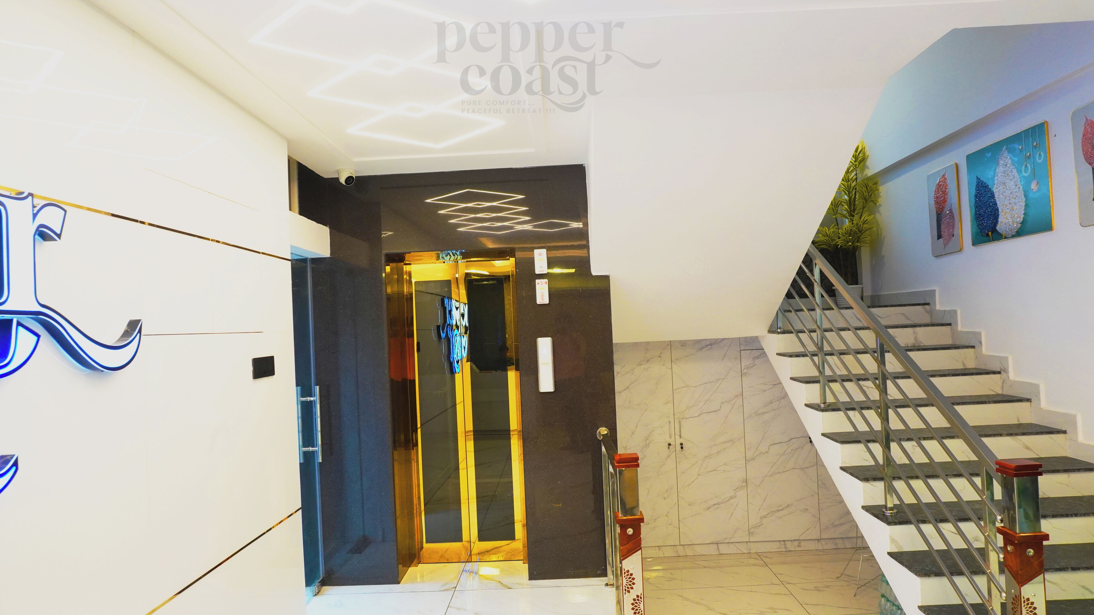 A modern hotel lobby with a marble staircase, a glass elevator, and a wall-mounted sign.