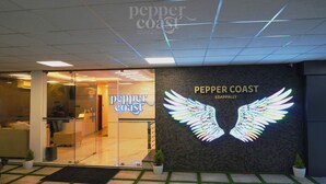 Exterior del Pepper Coast Edappally, con un letrero de neón que muestra un logotipo alado, entrada de vidrio y plantas en macetas.