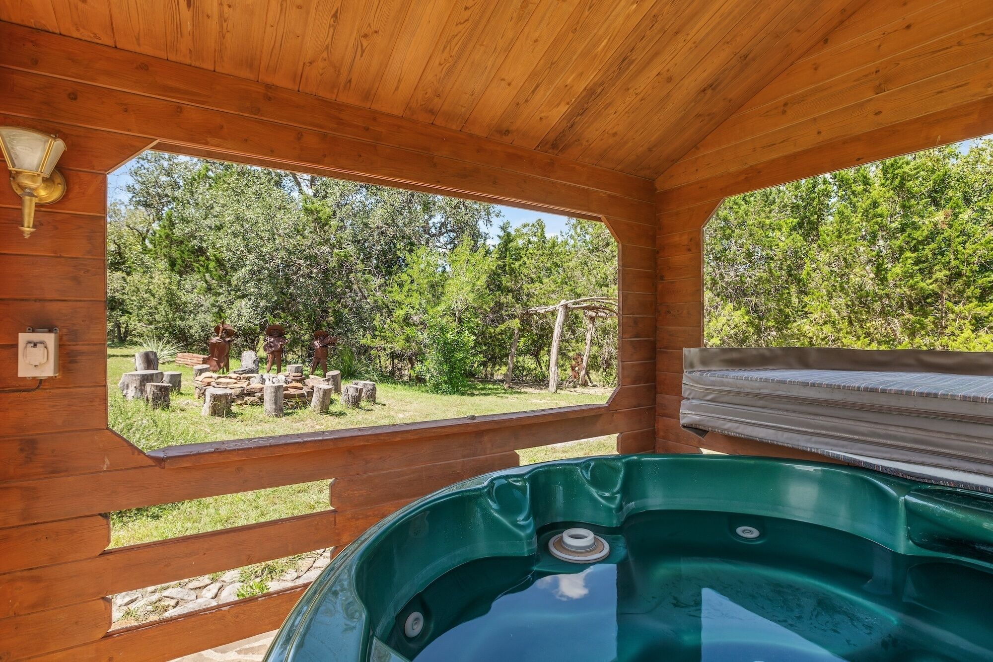 A wooden cabin with a hot tub, wooden ceiling, and a view of a garden with trees and a barbecue area.