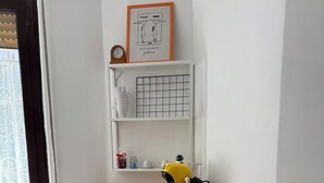 Une pièce de taille modeste, comprenant une table pliante, deux chaises, une étagère avec quelques objets et une fenêtre équipée de stores.