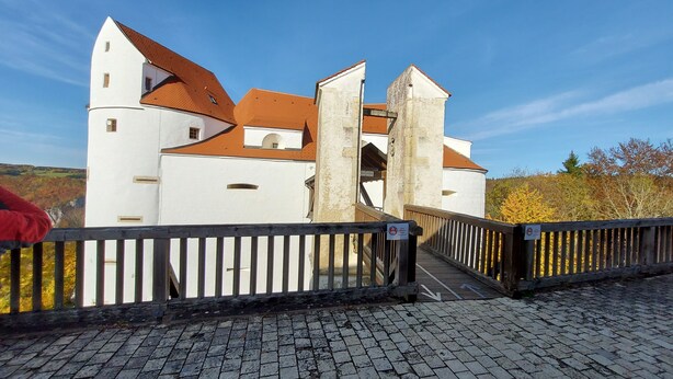 Un bâtiment historique à la façade blanche et au toit recouvert de tuiles rouges, un pont en bois menant à l’entrée, et un chemin pavé.