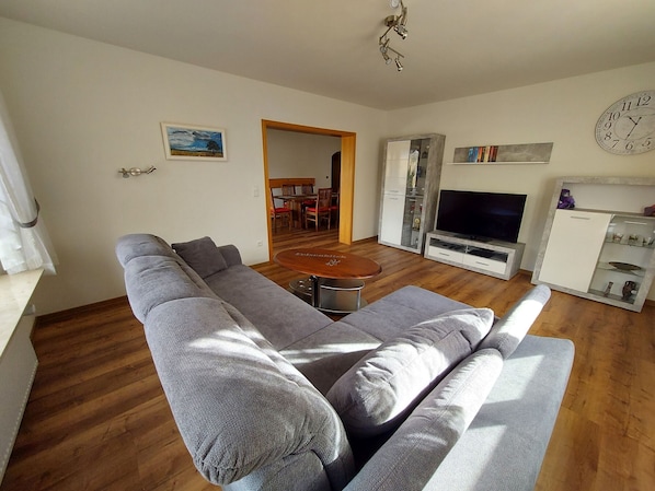 A living room with a grey sofa, wooden flooring, a TV, a clock, and a dining area in the background.