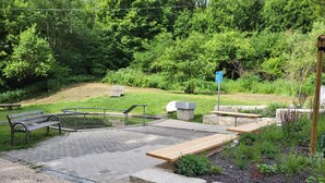 Un parc doté d’une zone dallée, de bancs, d’un petit élément décoratif aquatique et d’une végétation luxuriante.