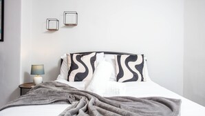 Een net opgemaakt bed met een grijze deken, een bedlampje en twee aan de muur bevestigde fotolijstjes.