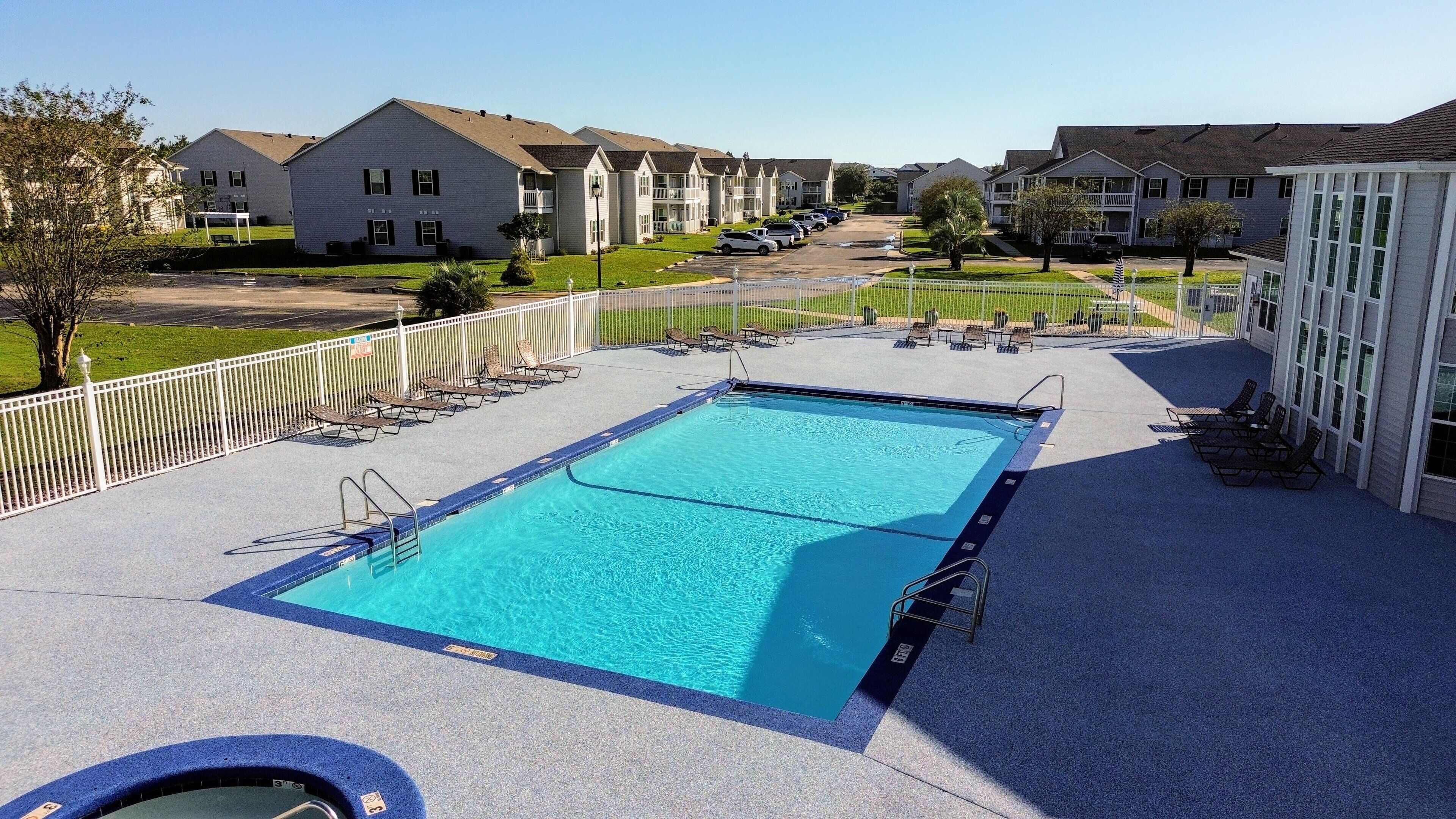 A rectangular swimming pool with a blue border, surrounded by a white fence. There are lounge chairs and a small pool table on the poolside.