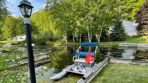 En brygga med en liten båt och en lyktstolpe vid en stilla damm, omgiven av grönska.