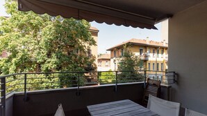 Un balcón con mesa y sillas de madera, baranda de metal y vista a edificios y árboles.