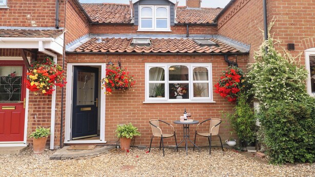 A brick house with a tiled roof, a front door, and two windows. There are potted plants and a small table with chairs outside. - Honeysuckle Cottage (Swaffham)