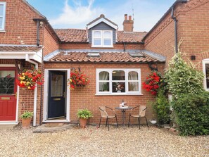 A brick house with a tiled roof, a front door, and two windows. There are potted plants and a small table with chairs outside.