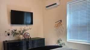 Een slaapkamer met een bed, een dressoir, een televisie, een airconditioner en een raam met jaloezieën.