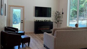 Een moderne woonkamer met een open haard, een flatscreen televisie en een groot raam met uitzicht op bomen.