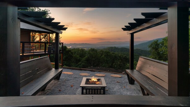 Una terraza de madera con un hogar y bancos, con vistas a un paisaje montañoso y arbolado al atardecer.