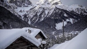 A wooden cabin with a snow-covered roof, surrounded by snow-covered mountains and trees.