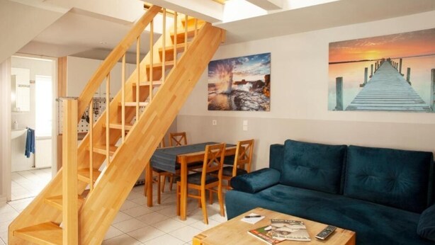 A modern living room with a wooden staircase, a blue sofa, a dining table with chairs, and two paintings on the wall. - Altstadtfreude Stralsund 5 (Stralsund)