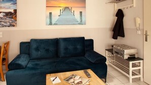 A living room with a dark blue sofa, a wooden coffee table with a magazine, a painting of a pier, and a coat rack with a jacket. - Altstadtfreude Stralsund 5 (Stralsund)