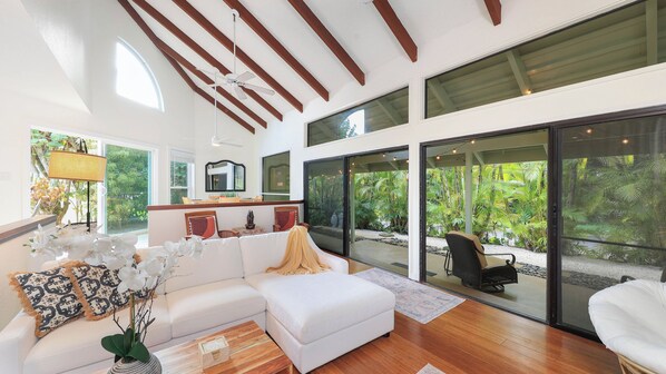 A spacious living room with a white sofa, a coffee table, and a view of a lush garden through large sliding glass doors. - Hale H Omaka in Princeville (Princeville)