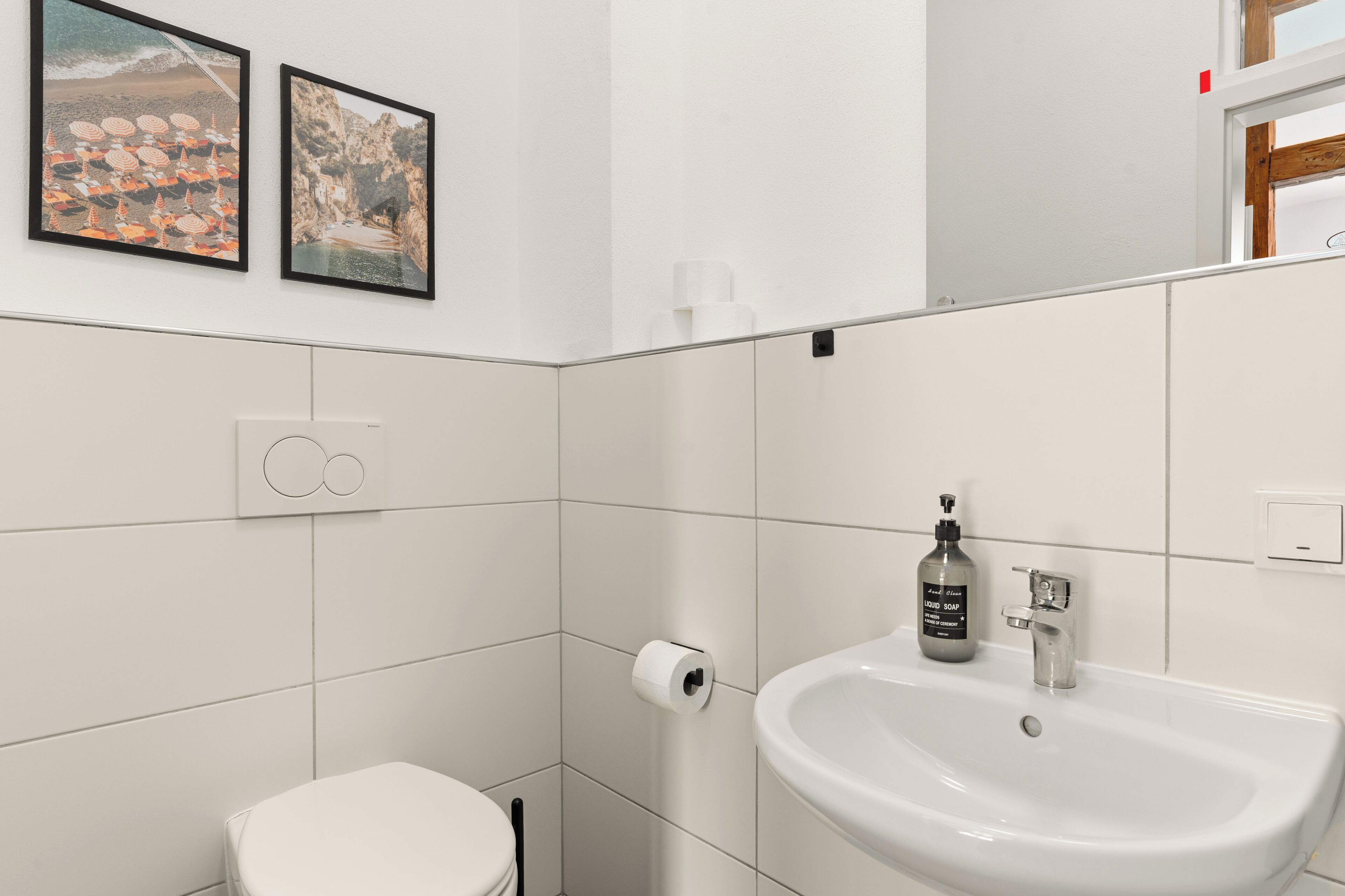 A modern bathroom with a white sink, toilet, and tiled walls. There are two framed pictures on the wall.