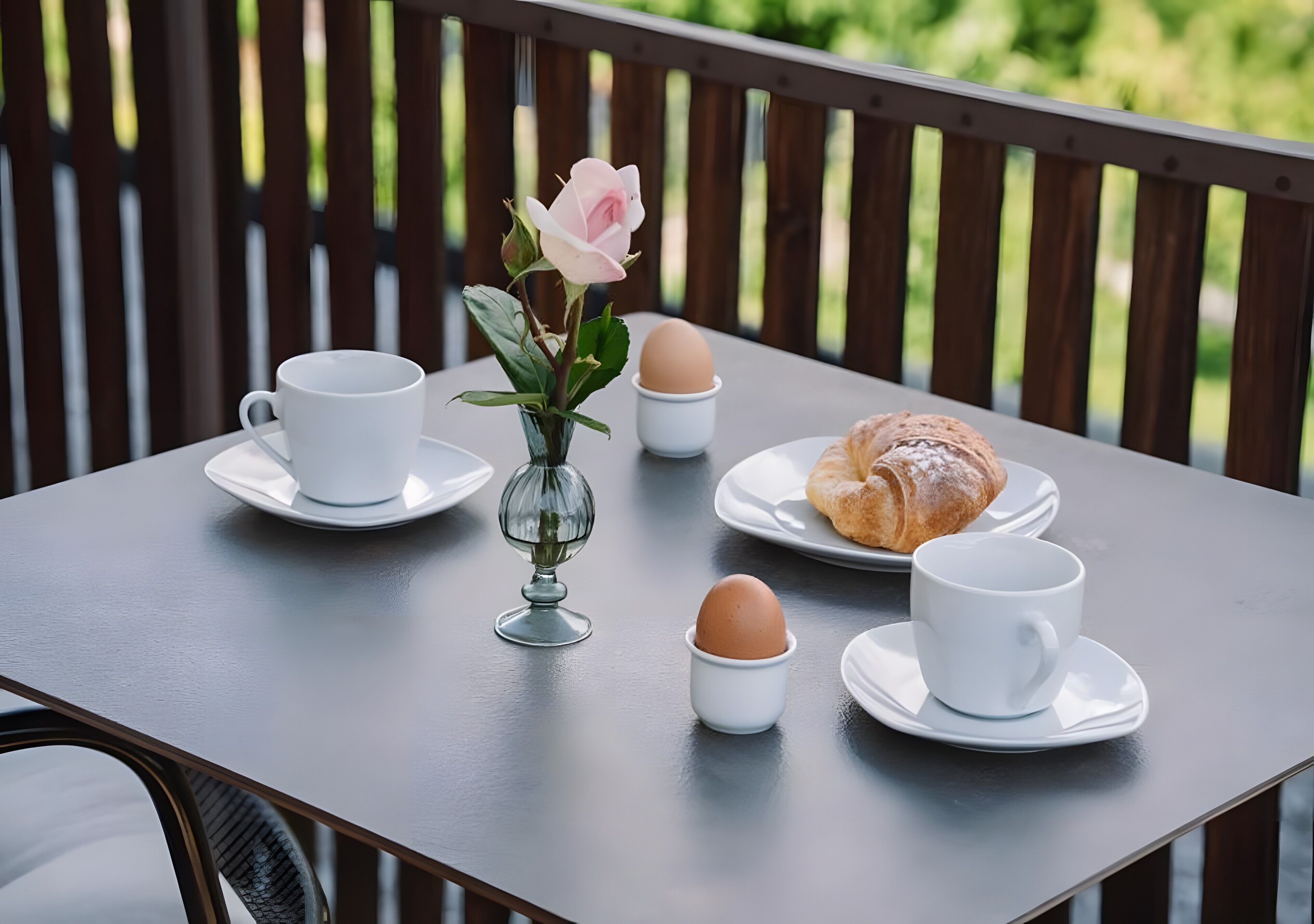 Ein Tisch mit einer weißen Tasse, einer Glasvase mit einer rosa Blume, einem Croissant und zwei Eiern auf Untertellern.