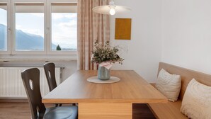 A dining area with a wooden table, chairs, a vase with flowers, a pendant light, and a view of mountains through a window.