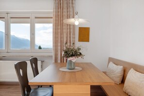 Un comedor con una mesa de madera, sillas, un florero con flores, una lámpara colgante y vistas a montañas a través de una ventana.