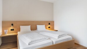 A modern hotel room with a wooden headboard, two beds with white bedding, bedside tables with lamps, and a white wall.