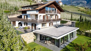 A modern two-story house with a balcony, a patio with outdoor furniture, and a view of a mountainous landscape.