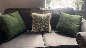 A grey sofa with three green cushions, one with a leaf pattern, and a window with a curtain and a vase with plants. - Sherwood Hideaway Retreat (Nottingham)