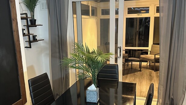 Een eethoek met een zwarte tafel, zes stoelen, een potplant en een groot raam met gordijnen.